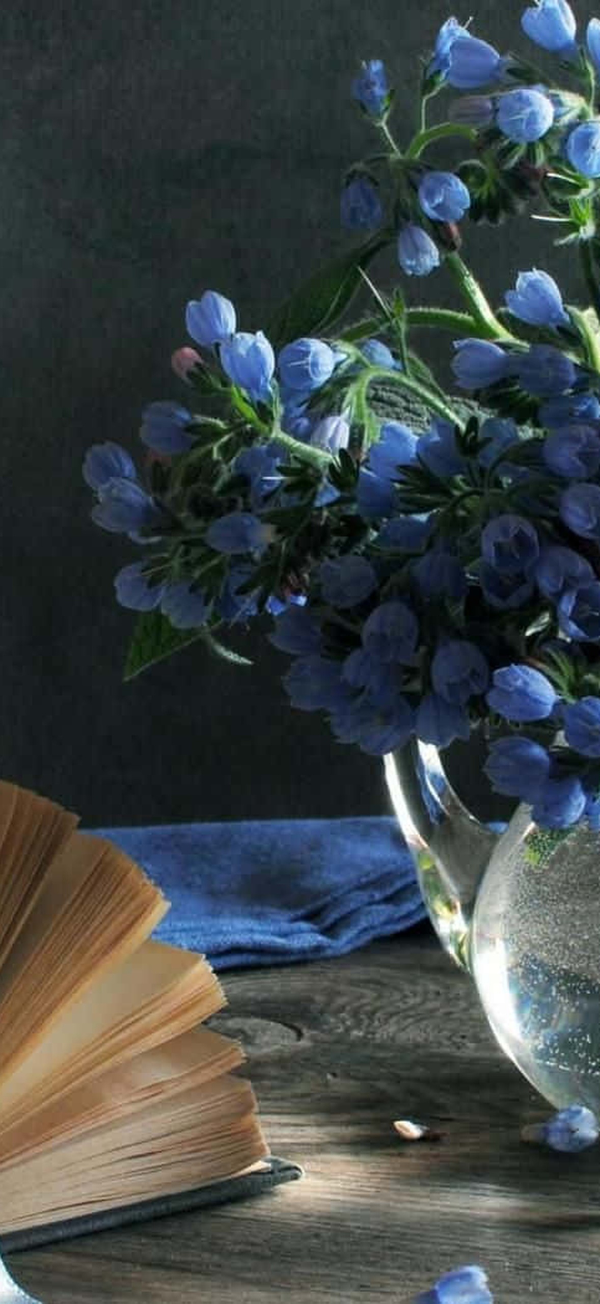 Blue borage flowers in a glass vase next to an open book on wood.