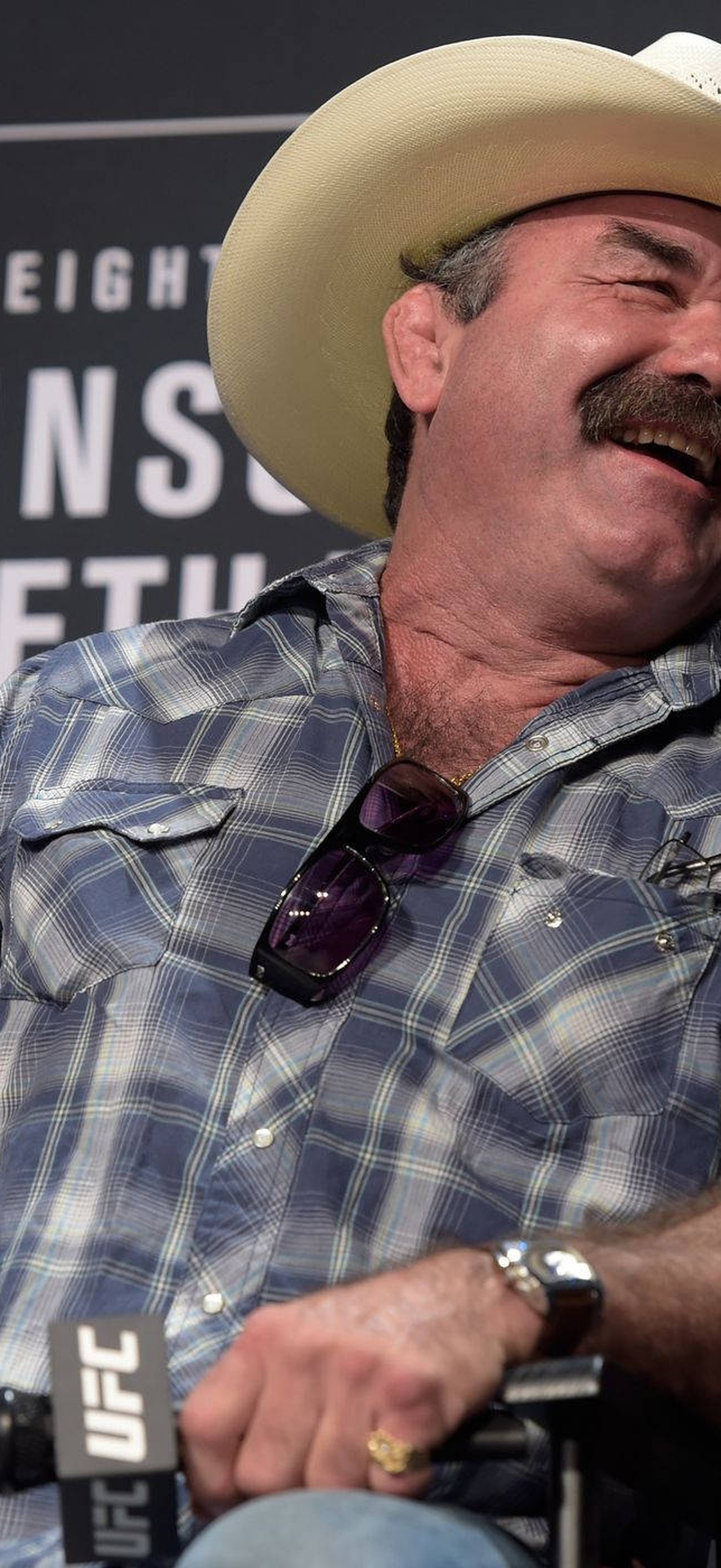 Man in cowboy hat and plaid shirt laughing at UFC press event.