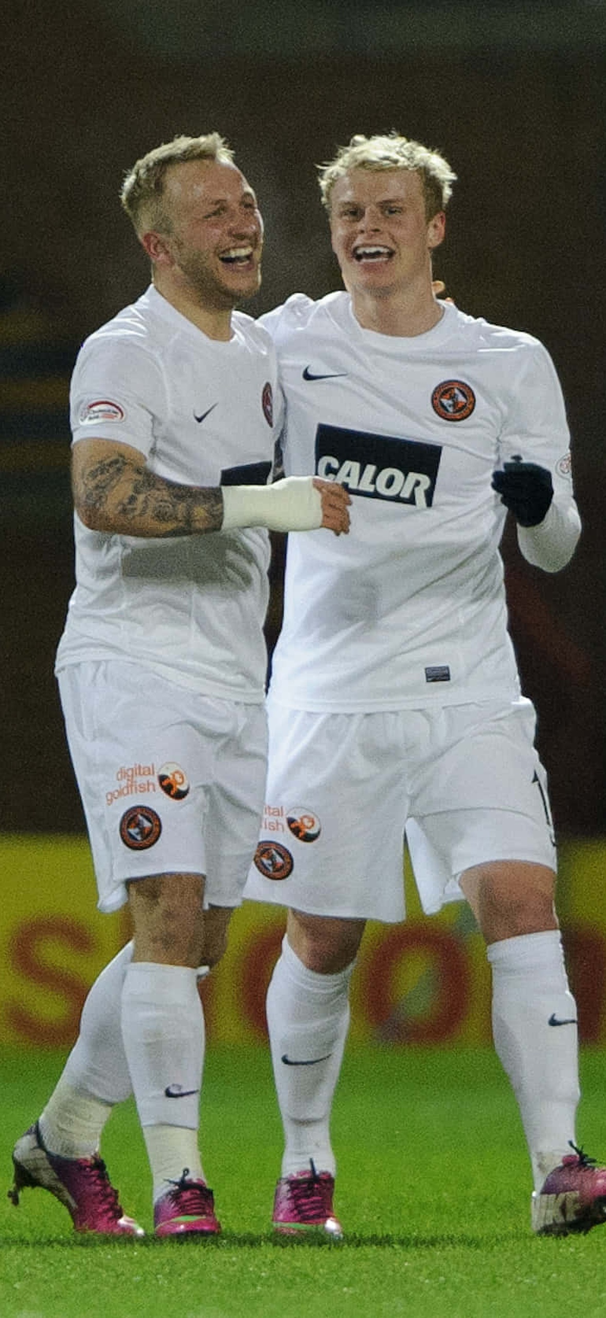 Two soccer players laughing and celebrating a goal on a lit field.
