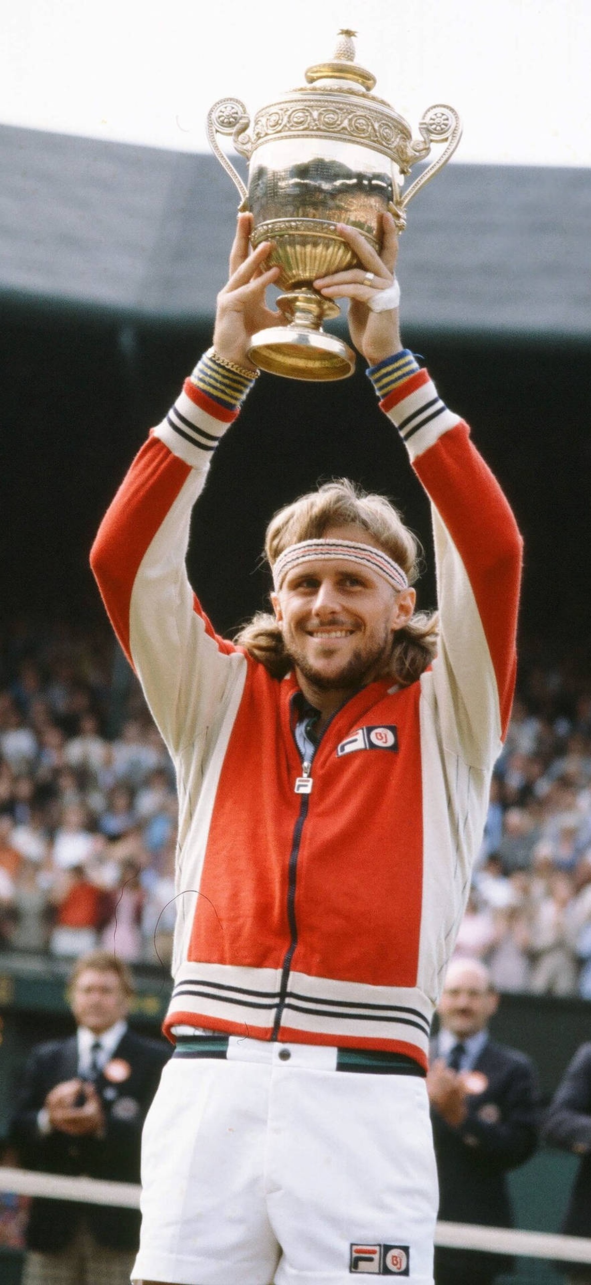 Tennis champion celebrates victory by holding up a large silver trophy on grass court.