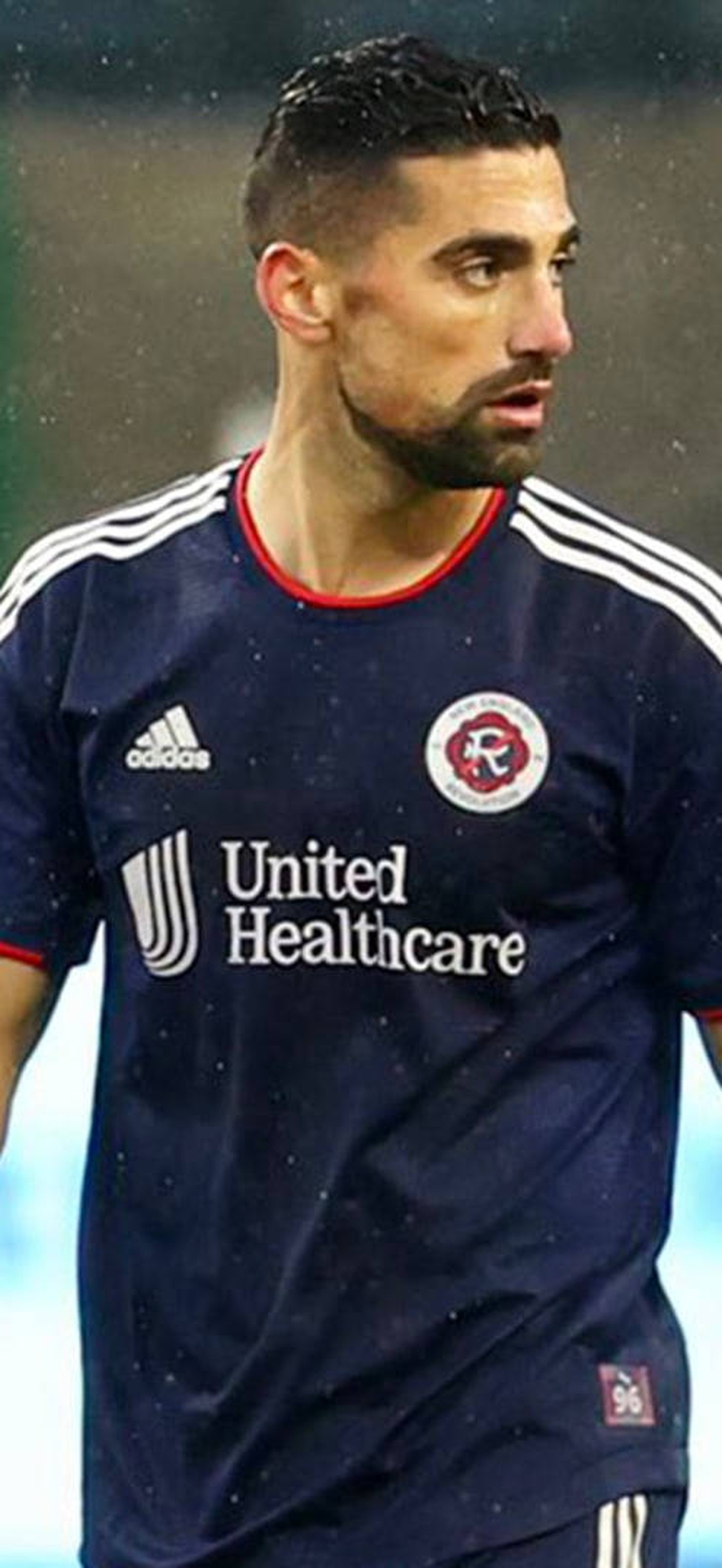 Soccer player in navy jersey walking on rainy field at night.