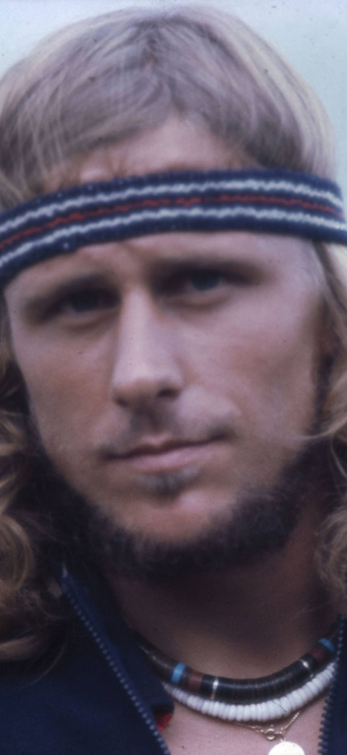 Man with long blonde hair and striped headband wearing a beaded necklace.